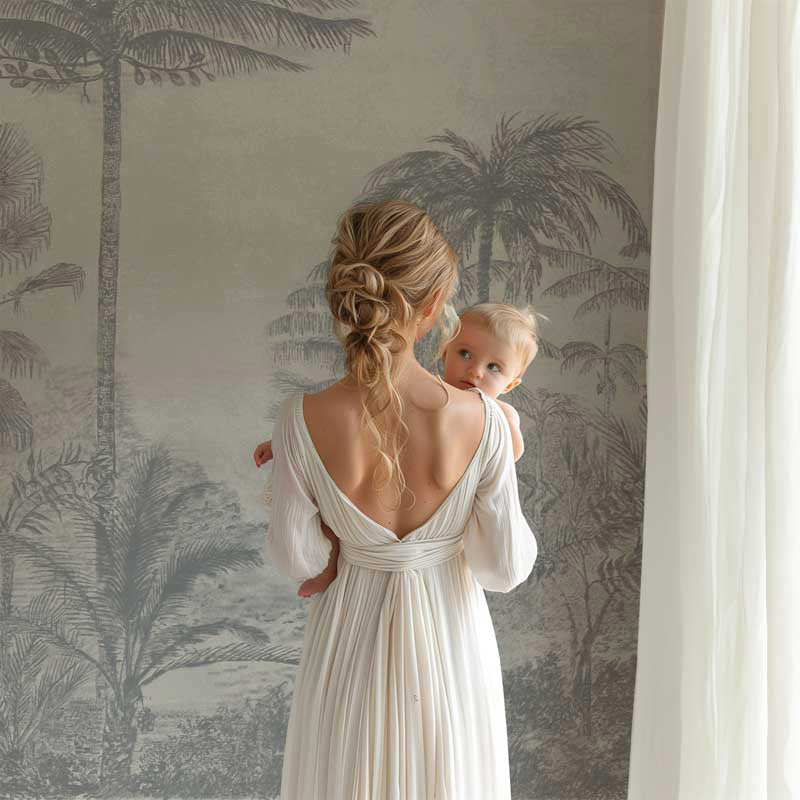 Woman in a white dress holding a baby against a palm tree wallpaper wall.