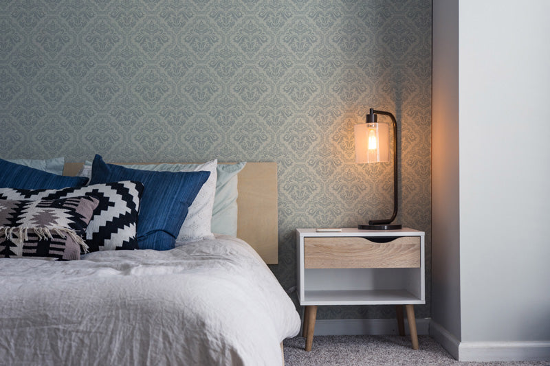 Bedroom with damask wallpaper, bed with pillows and blanket, and nightstand with lamp.