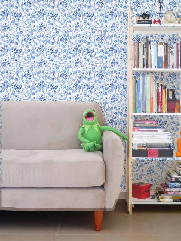 Green frog plush toy on a beige sofa against a blue floral wallpaper with a bookshelf in the background.