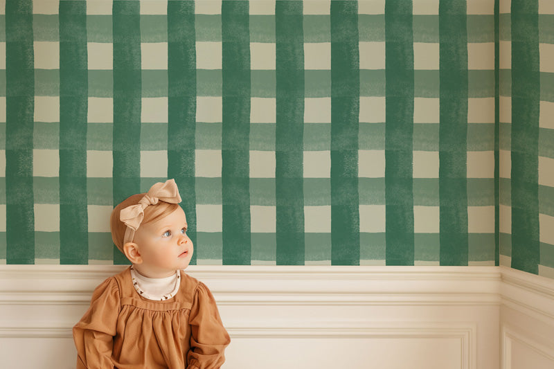 Baby in a brown dress standing in front of green and white checkered wallpaper